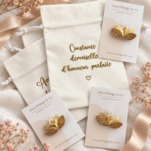 Op de afbeelding: Drie goudkleurige bloemvormige broches met glitterende bloemblaadjes bevestigd op witte displaykaarten met het label Sweethings & co. De broches liggen bij crèmekleurige stoffen trekkoordzakjes, waarvan er één de tekst Constance demoiselle d'honneur parfaite met een klein hartje bevat.