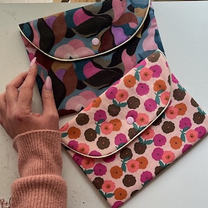 May include: Two fabric pouches with rounded flaps. One pouch has a floral pattern in pink, orange, and brown on a pink background. The other pouch has a pattern of abstract shapes in shades of blue, pink, and brown. Both have a light-colored trim and a pink button closure.