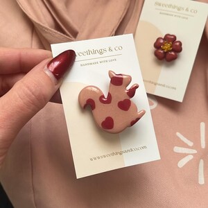 May include: A pink ceramic squirrel pin with red hearts on it. The pin is on a white card with the text "Sweethings & Co Handmade with Love" and the website "www.sweethingsandco.com".