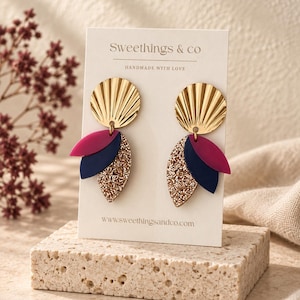 Peut inclure: Une paire de boucles d'oreilles pendantes sur un support marqué Sweethings & co. Chaque boucle présente un disque doré plissé surmontant trois éléments en forme de feuille superposés, de couleur magenta, bleu marine et pailleté or.