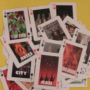 May include: A close-up of a pile of playing cards with various images and numbers. The cards are arranged in a random order, with some cards partially visible. The cards feature images of a four-leaf clover, roses, a city skyline, the Magi, and a succubus.