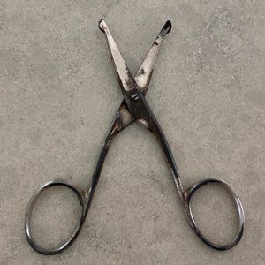 May include: A pair of vintage metal scissors with rounded finger holes and pointed blades. The scissors are silver-toned with signs of wear. The blades are crossed and held together by a screw.