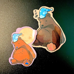May include: Two brown bear stickers with blue sleeping caps. One bear is sitting with a pink paw and the other is sitting with a blue paw. The bear on the left has a yellow sun behind it.