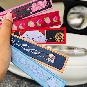 May include: A collection of embroidered keychains in various colors. Designs include pink roses, strawberries, a white cat, a cartoon character, and a seal. Each keychain has a different color scheme and design, with a metal ring for attachment.