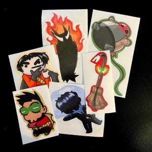 Puede incluir: Una colección de seis pegatinas troqueladas con ilustraciones estilo dibujos animados de varios personajes y objetos, incluyendo una figura en llamas, un personaje bebiendo café, un héroe con máscara verde, una figura oscura y una palanca.