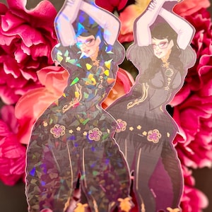 May include: Two holographic stickers of a woman in a black dress holding a gun. The woman has black hair and is wearing glasses. The stickers are on a pink background.