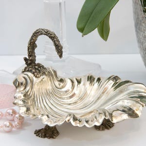 Peut inclure: Un plat décoratif argenté avec un motif en forme de vague et une poignée incurvée. Le plat a une surface texturée et détaillée et repose sur de petits pieds ornés. Un ruban rose et un collier de perles sont au premier plan.