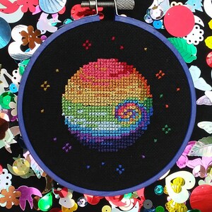 May include: A rainbow-coloured cross-stitch design of a spiral galaxy, surrounded by colourful sequins and beads. The design is stitched on a black fabric and framed in a dark blue embroidery hoop.
