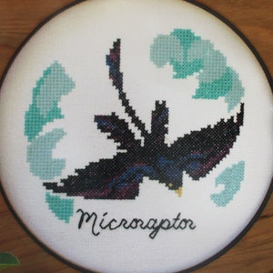 May include: A framed cross-stitch embroidery of a Microraptor, a small feathered dinosaur, with a black and purple body and green leaves in the background.
