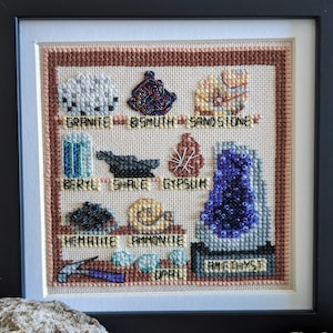 Könnte beinhalten: Ein gerahmter Kreuzstich-Sampler mit verschiedenen Mineralien und Edelsteinen, darunter Granit, Beryll, Hämatit und Amethyst. Jedes Mineral ist mit seinem Namen beschriftet.