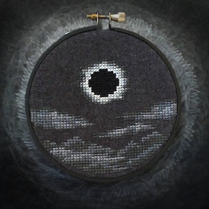 May include: A black and white cross-stitch embroidery of a solar eclipse with a dark circle in the center, surrounded by a white ring and a dark gray background. The embroidery is framed in a black hoop with a gold screw.