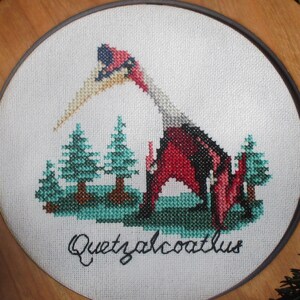 May include: A framed cross-stitch embroidery of a red and black Quetzalcoatlus dinosaur with a long beak, surrounded by green trees. The embroidery is titled "Quetzalcoatlus".