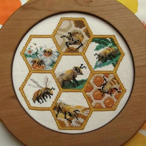 May include: A framed cross-stitch design depicting a honeycomb with bees and flowers. The honeycomb is made up of six hexagonal cells, each with a different scene. The cells feature bees, flowers, and a honeycomb pattern.