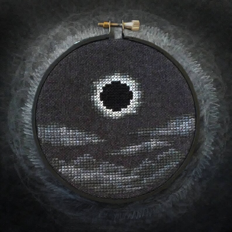 May include: A black and white cross-stitch embroidery of a solar eclipse with a dark circle in the center, surrounded by a white ring and a dark gray background. The embroidery is framed in a black hoop with a gold screw.