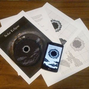 May include: A close-up of a black and white cross-stitch pattern of a solar eclipse. The pattern is displayed on a smartphone screen and a printed sheet of paper. The text "Solar Eclipse" is printed on the paper.