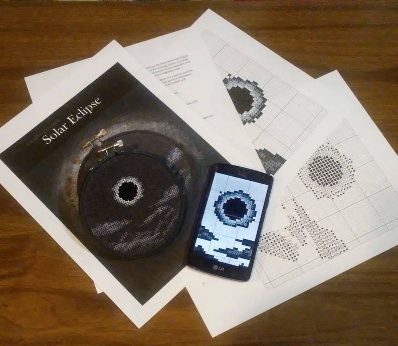 May include: A close-up of a black and white cross-stitch pattern of a solar eclipse. The pattern is displayed on a smartphone screen and a printed sheet of paper. The text "Solar Eclipse" is printed on the paper.