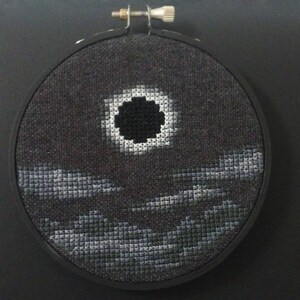 May include: A black and white cross-stitch embroidery of a dark circle with a white ring around it, set against a dark gray background with three rows of gray clouds.