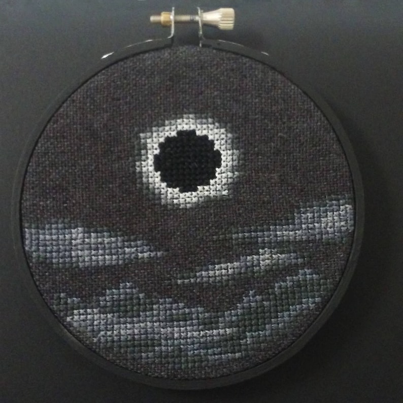 May include: A black and white cross-stitch embroidery of a dark circle with a white ring around it, set against a dark gray background with three rows of gray clouds.