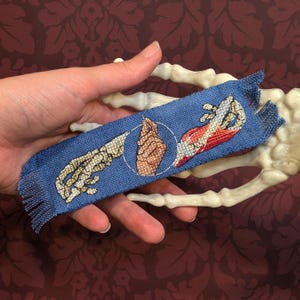 May include: A blue bookmark with a fringed edge, featuring embroidered anatomical illustrations of hands and a section of muscle. The design includes a skeletal hand, a flesh-toned hand, and a red and white muscle section. The bookmark is held in a hand.