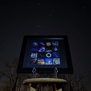 May include: A framed cross-stitch piece depicting a grid of space-themed images, including planets, stars, and a rocket ship. The frame is black and the cross-stitch is in shades of blue, purple, and white.