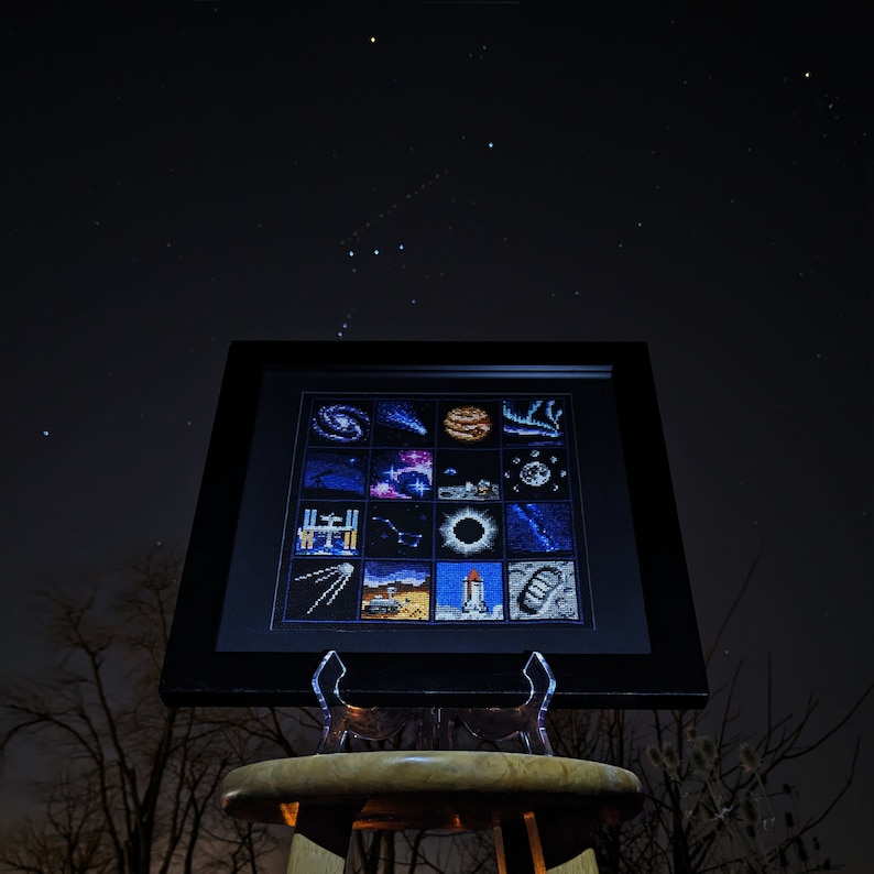 May include: A framed cross-stitch piece depicting a grid of space-themed images, including planets, stars, and a rocket ship. The frame is black and the cross-stitch is in shades of blue, purple, and white.