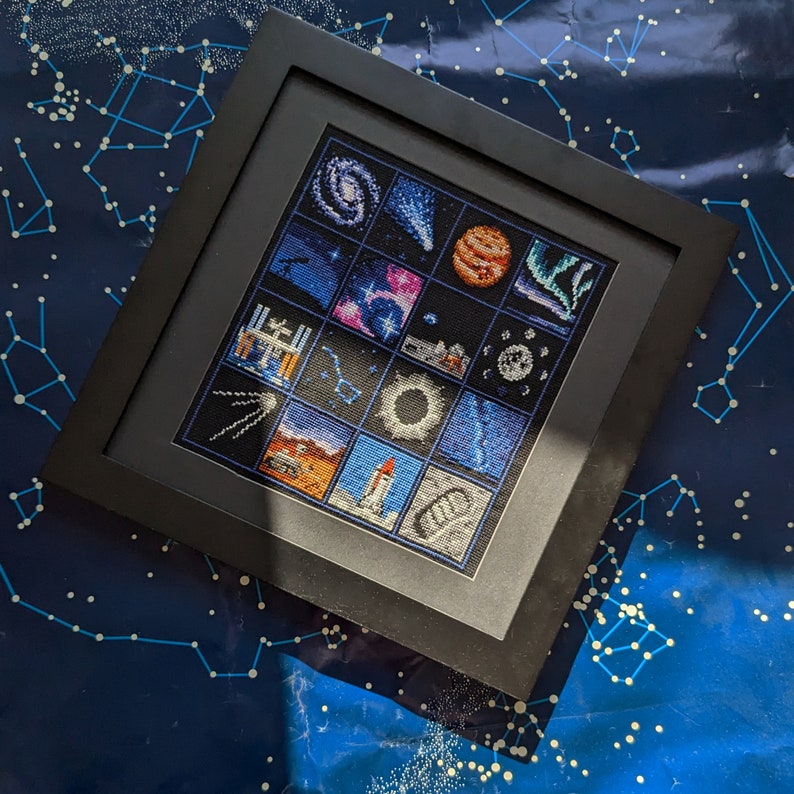 May include: A black framed cross-stitch with a space theme. The cross-stitch features a variety of space-related images, including planets, stars, rockets, and the International Space Station.