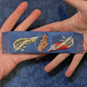 May include: A blue fabric bookmark with a cross-stitched design depicting two hands reaching towards each other, one skeletal and the other flesh-toned, within a white circle. The bookmark has fringed edges.