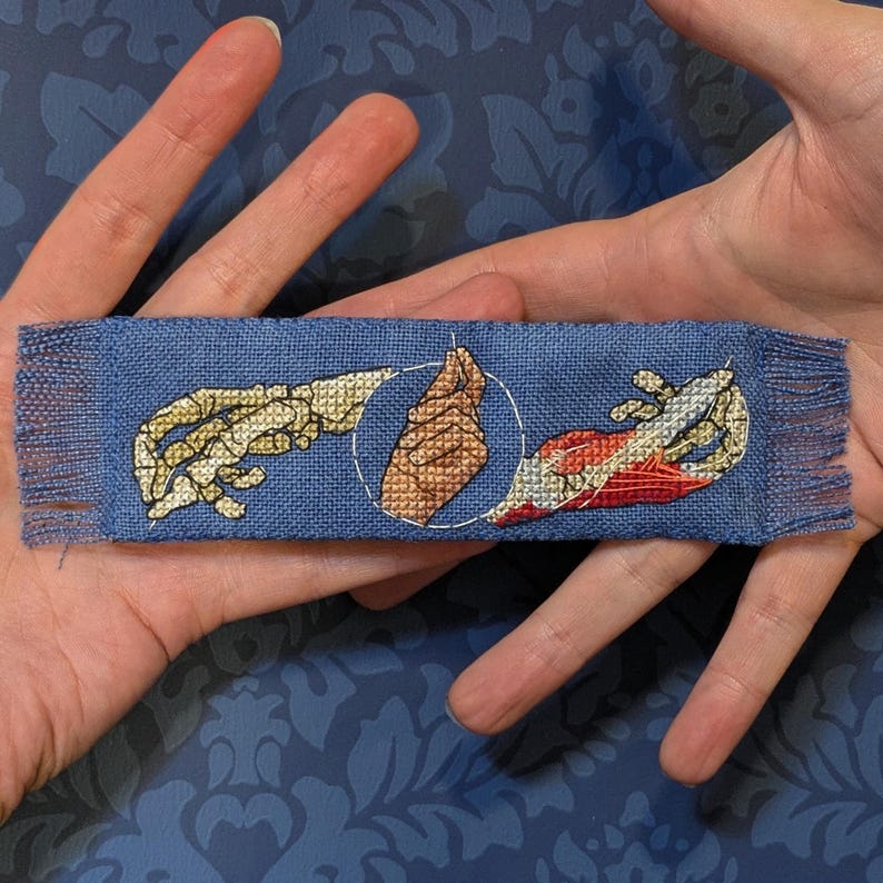 May include: A blue fabric bookmark with a cross-stitched design depicting two hands reaching towards each other, one skeletal and the other flesh-toned, within a white circle. The bookmark has fringed edges.