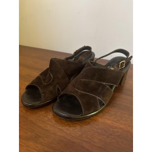 May include: A pair of brown suede sandals with open toes and slingback straps. The sandals have a block heel and gold-colored buckles. The shoes are displayed on a wooden surface.