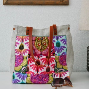 Könnte beinhalten: Eine große Tragetasche mit einem Blumenmuster in Rosa, Lila und Gelb. Die Tasche hat braune Ledergriffe und eine hellgraue Leinenaußenseite. Eine Sonnenbrille liegt vor der Tasche.