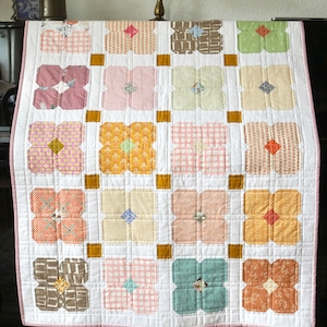 Op de afbeelding: Een handgemaakte quilt met een bloemenpatroon. De quilt heeft een witte achtergrond met een raster van kleurrijke bloemvormige blokken. Elk blok heeft een andere stof met patronen in tinten roze, geel, groen en bruin. De quilt is afgezet met een rood-wit gestreepte stof.