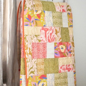 Könnte beinhalten: Ein Patchwork-Quilt mit verschiedenen floralen und geometrischen Mustern in Rosa, Grün, Gelb und Weiß. Der Quilt hat einen orangefarbenen Rand und hängt in der Nähe eines Fensters mit transparenten Vorhängen.
