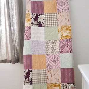 Könnte beinhalten: Ein Patchwork-Quilt mit quadratischen Mustern in Lila, Gold und Grün. Der Quilt hat florale, geometrische und karierte Designs. Der Quilt hängt an einer weißen Wand in der Nähe eines Fensters mit einem grauen Vorhang.