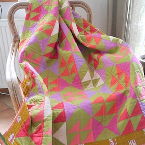 Peut inclure: Une courtepointe colorée avec un motif géométrique, drapée sur une chaise en osier. La courtepointe présente des triangles verts, roses, violets et blancs. Les bords sont finis avec une bordure contrastante.