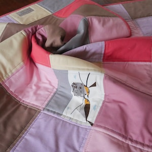 Könnte beinhalten: Ein Patchwork-Quilt in Rosa-, Braun-, Grau- und Cremetönen. Der Quilt weist ein sich wiederholendes quadratisches Muster mit einem skurrilen Vogeldesign auf einem Abschnitt auf. Der Quilt ist gefaltet und zeigt die verschiedenen Farben und Texturen.