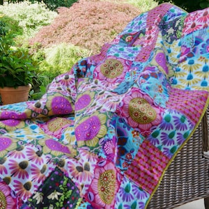 Könnte beinhalten: Eine bunte Patchwork-Decke mit floralen Mustern, drapiert über einem Korbstuhl. Die Decke zeigt große Blumen in Rosa, Lila, Blau und Grün. Sie hat einen gelben Rand, der eine lebendige und gemütliche Ästhetik erzeugt.