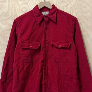 Puede incluir: Una camisa roja de manga larga con dos bolsillos en el pecho y cierre de botones. La camisa está colgada en una percha de madera sobre un fondo claro de vetas de madera. La camisa tiene un estilo clásico e informal.