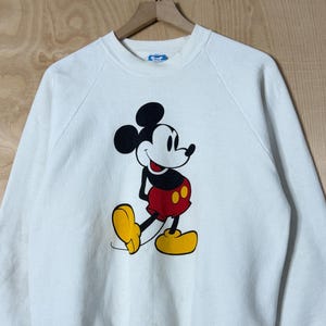May include: White sweatshirt featuring a large graphic of Mickey Mouse. The cartoon character is depicted in his classic pose, with black ears, a red and yellow outfit, and yellow shoes. The sweatshirt has long sleeves and a crew neck.