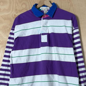 Op de afbeelding: Een rugby shirt met lange mouwen en horizontale strepen in paars, wit en groen. Het shirt heeft een blauwe kraag en manchetten, en een kleine logopatch bij de zoom. Het hangt aan een houten hanger.