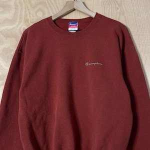 Op de afbeelding: Een bordeauxrood Champion sweatshirt met een ronde hals en lange mouwen. Het Champion-logo is in het wit op de borst geborduurd. De sweatshirt heeft geribde manchetten en zoom. Het kledingstuk hangt aan een houten kleerhanger.