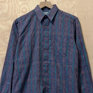May include: A long-sleeved button-down shirt with a purple and teal vertical stripe pattern. The shirt has a collared neckline, a chest pocket, and is hanging on a wooden hanger.