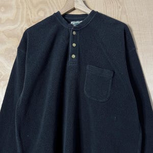 May include: A black fleece pullover shirt with a Henley neckline, three buttons, and a single pocket. The long-sleeved shirt is hanging on a wooden hanger against a light wood grain background.