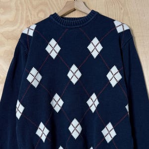 May include: Navy blue argyle sweater with white diamond shapes and maroon accent lines. The sweater has a ribbed crew neck, cuffs, and hem. The sweater is hanging on a wooden hanger against a light wood background.