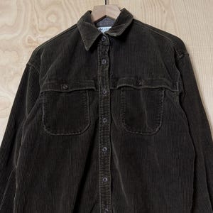 May include: A dark olive green long-sleeve button-down shirt made of corduroy fabric. The shirt features a standard collar, a full front button placket, and two chest pockets with buttoned flaps. The cuffs are also buttoned, and the hem is straight.