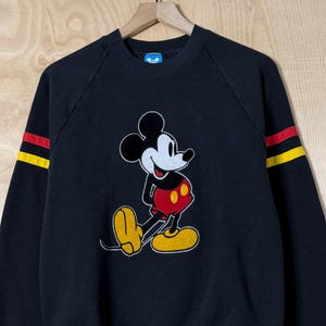 May include: Black sweatshirt featuring a classic Mickey Mouse graphic. The long sleeves have yellow and red stripes. The cartoon character is depicted in his signature outfit with yellow shoes, red shorts, and white gloves.