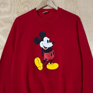 May include: Red sweatshirt with a Mickey Mouse graphic. The cartoon character is shown in his iconic outfit: black ears, white face, red shorts, yellow shoes, and white gloves. The long-sleeved top is suspended from a wooden hanger.
