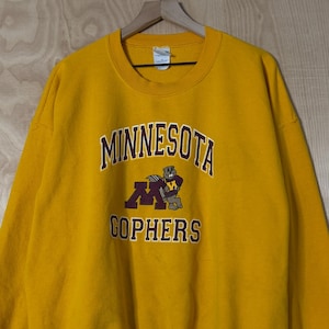 May include: Mustard yellow sweatshirt with the text "MINNESOTA GOPHERS" in maroon and gold. The sweatshirt has a crew neck and long sleeves. The graphic features a football player.
