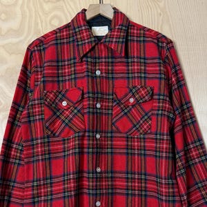 Puede incluir: Una camisa a cuadros roja y negra con dos bolsillos en el pecho. La camisa de manga larga está colgada de una percha de madera. El patrón de cuadros incluye líneas azul marino y amarillas.