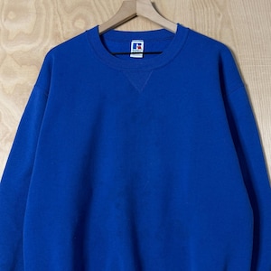 Puede incluir: Una sudadera azul real con cuello redondo y mangas largas. La sudadera tiene un pequeño logotipo blanco y azul en la manga inferior izquierda y una etiqueta en la parte superior del pecho. La sudadera está colgada de una percha de madera.