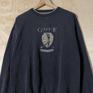 以下が含まれることがあります： チャコールグレーの長袖スウェットシャツ。クルーネックにはタンカラーのトリムが施されています。胸元には、ゴルファーが描かれた盾のグラフィックの上に「GOLF」、盾の下に「Classic Links」という文字が刺繍されたエンブレムがあしらわれています。